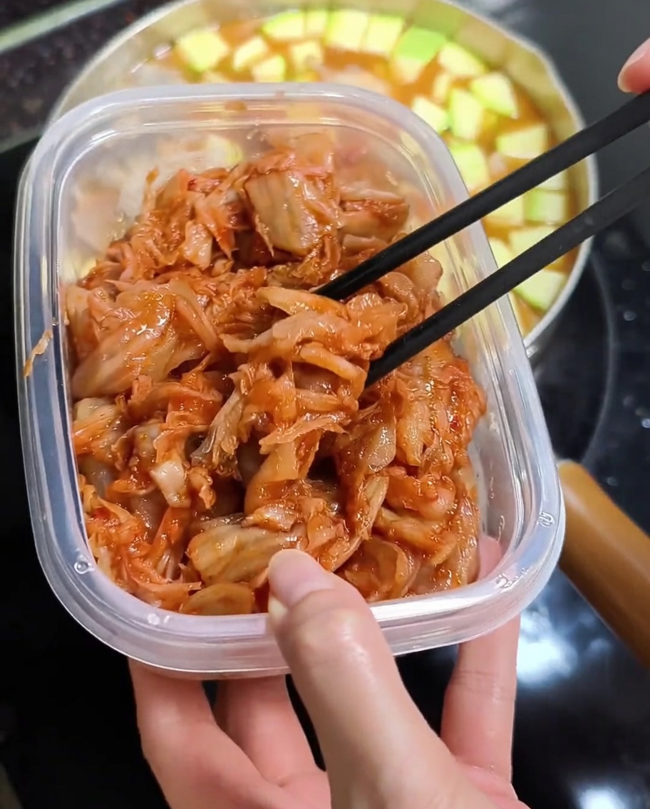 젓가락으로 플라스틱 용기에서 잘게 썬 붉은 김치를 집어 뜨거운 찌개 냄비 위로 올리는 모습.