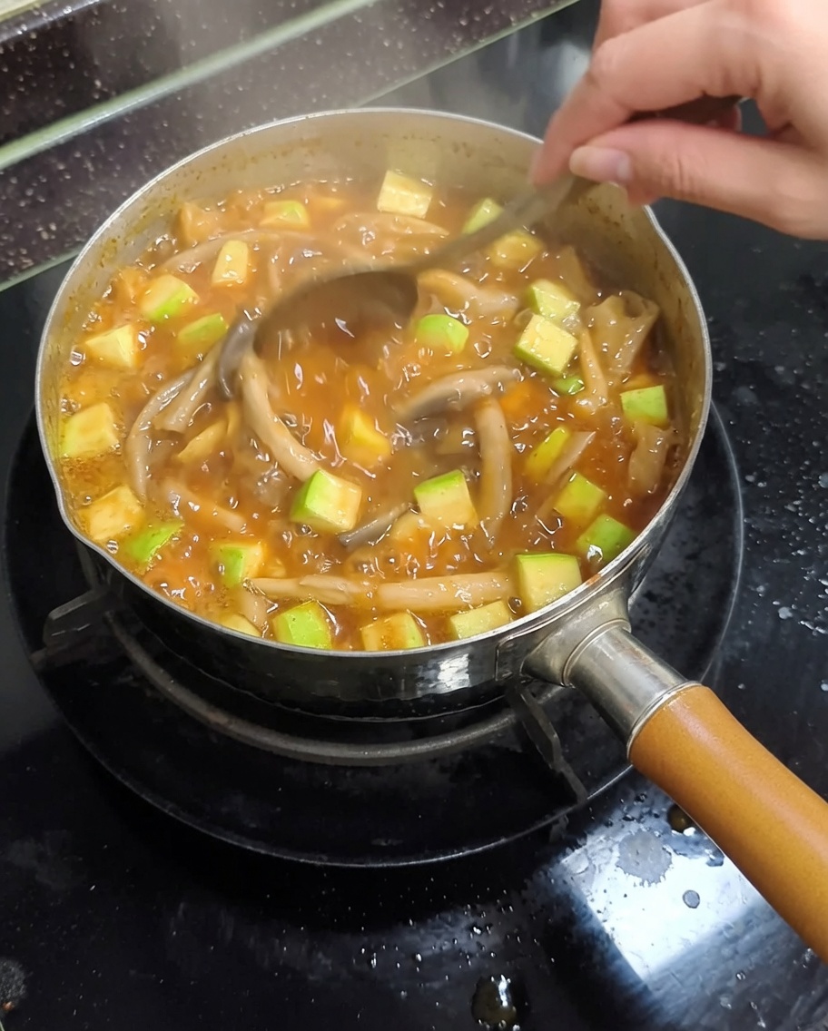 가스레인지 위에 있는 금속 냄비 안에서 된장찌개가 끓고 있고, 애호박과 버섯이 보이는 찌개를 금속 숟가락으로 젓는 모습.