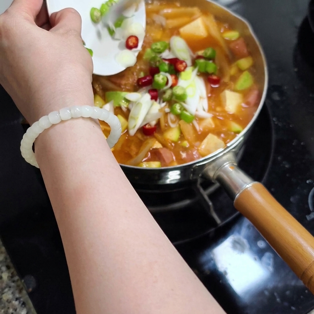 작은 흰 접시를 든 손이 썰어둔 청양고추, 홍고추, 대파를 끓고 있는 찌개 냄비에 붓는 모습.
