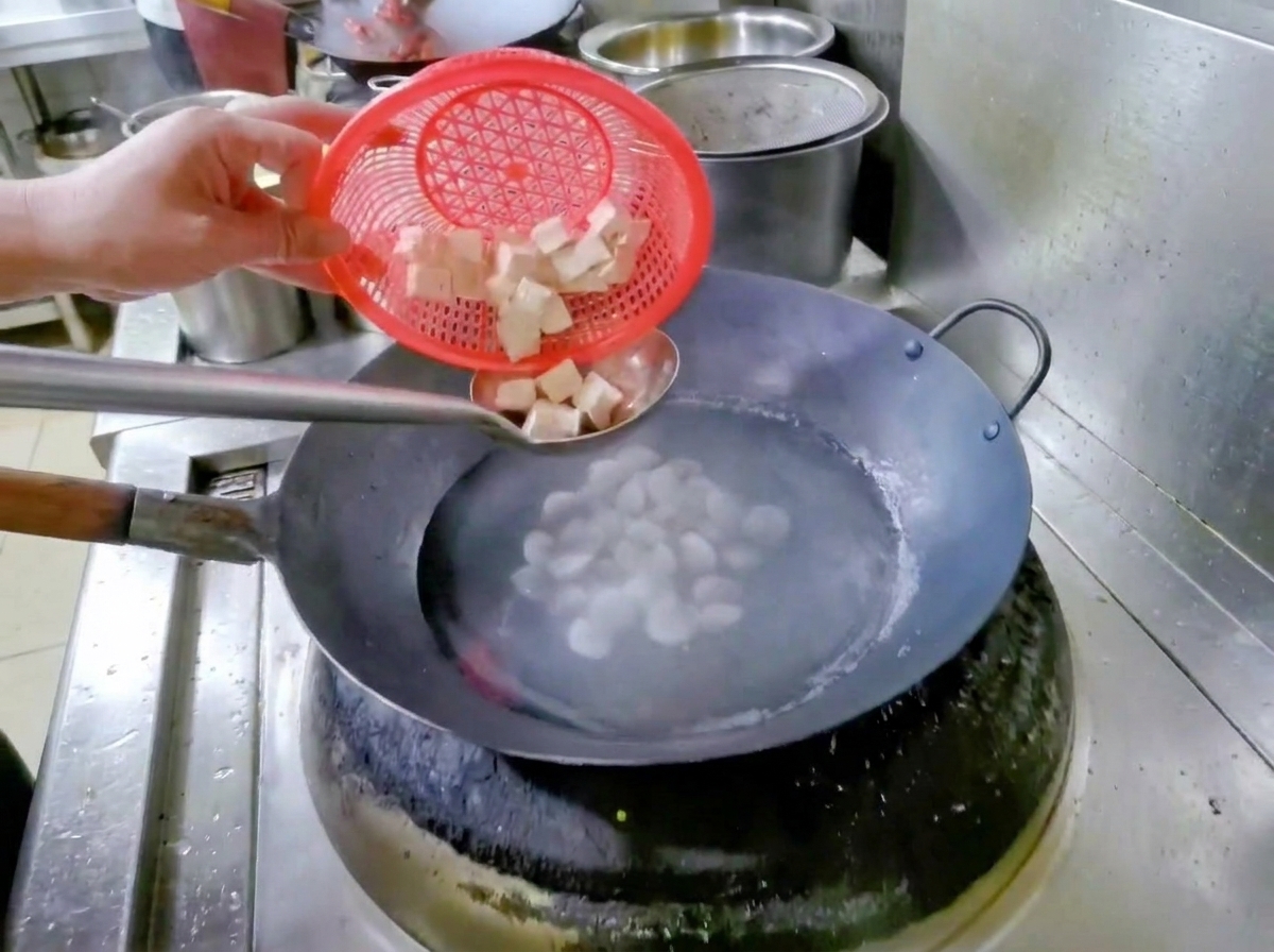 빨간색 거름망을 든 손이 물과 조개가 담긴 냄비에 깍둑 썰어 둔 두부를 넣고 있는 모습