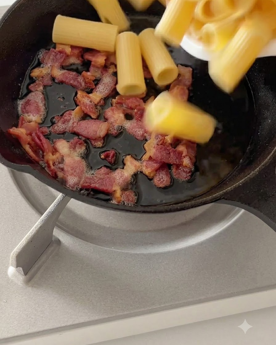 약불 위 팬에서 삶은 리가토니 파스타가 조리된 베이컨과 섞이는 모습.