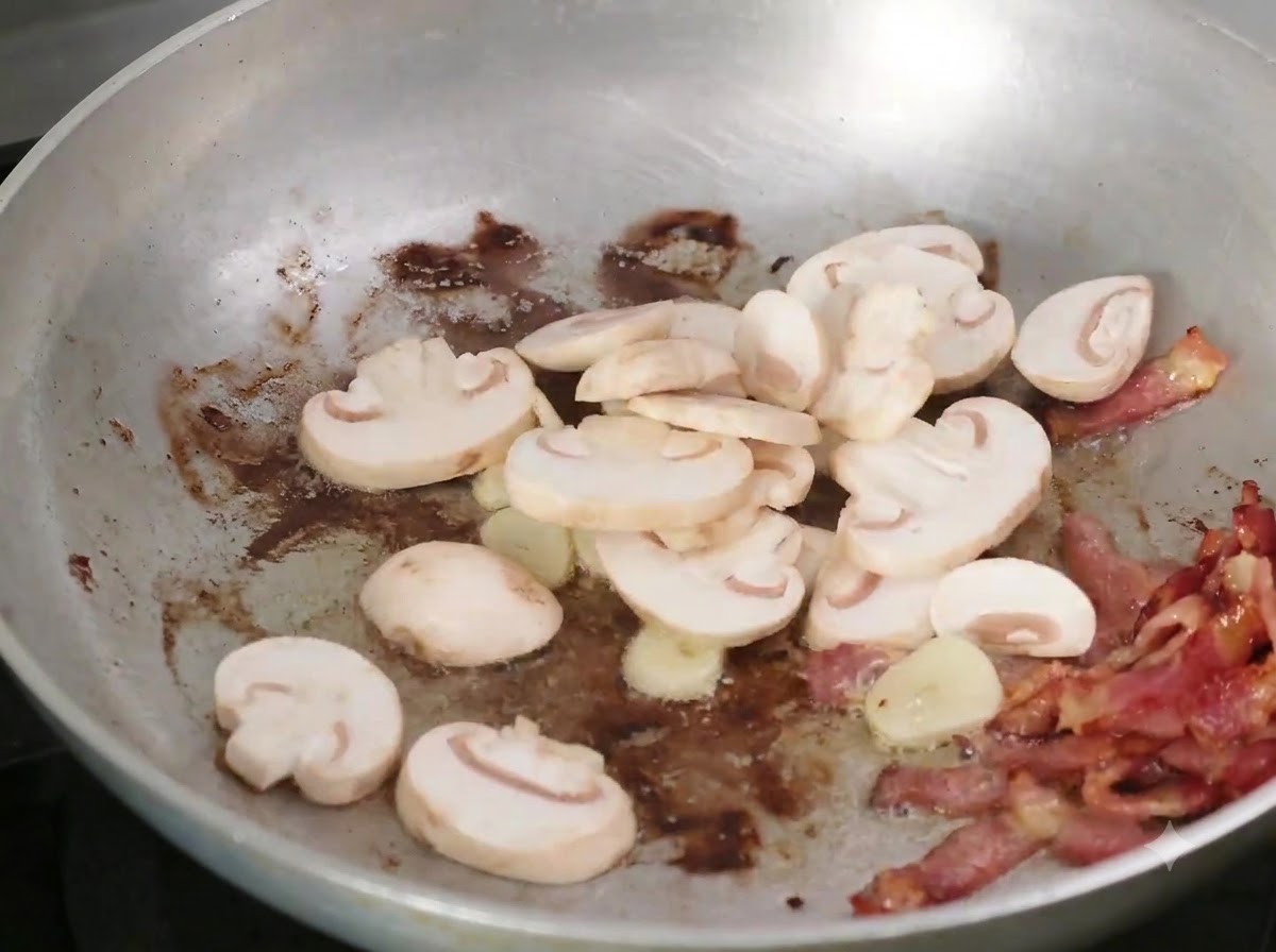 베이컨과 마늘이 있는 팬에 얇게 썬 버섯을 넣는 모습.