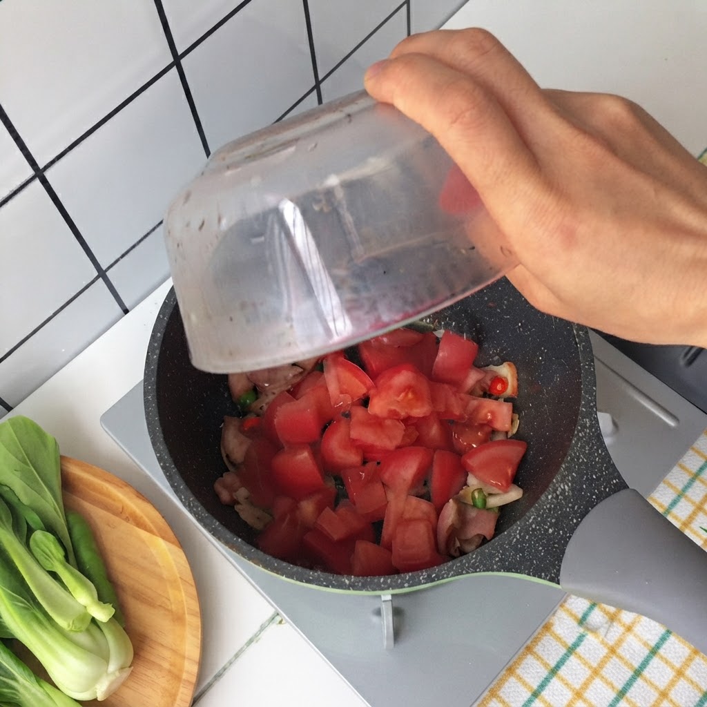 익은 베이컨과 대파가 담긴 팬에 유리 그릇에 든 썬 토마토를 붓고 있는 손.