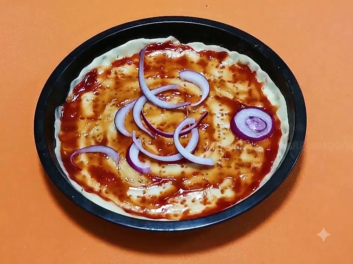 검은색 원형 베이킹 팬 안의 토마토 소스가 발린 피자 도우 위에 썰어놓은 붉은 양파 링이 배열된 모습.