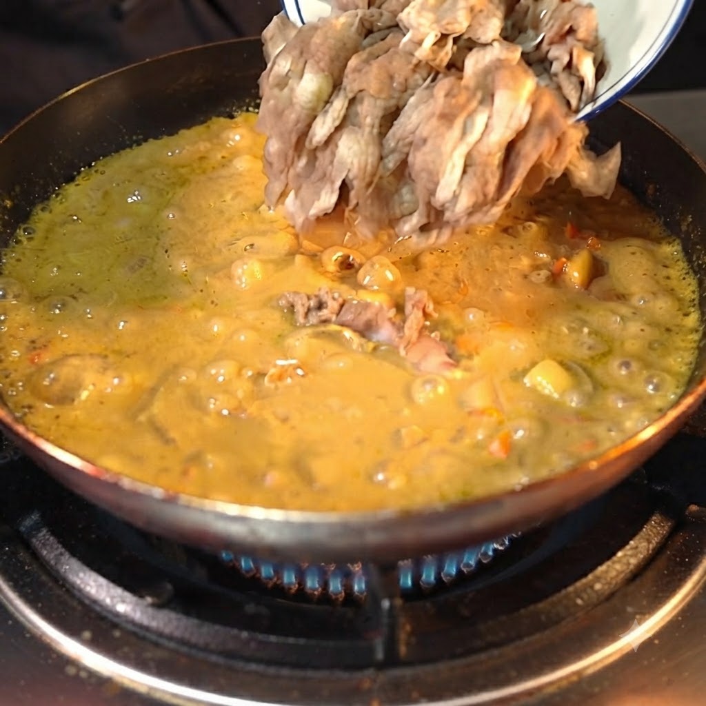 어두운 색상의 팬에서 걸쭉하게 끓고 있는 노란 카레 소스 속 흰색 그릇에서 부어지는 손질된 소고기 슬라이스.