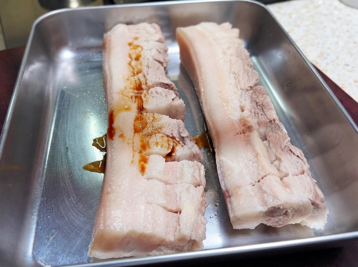 메탈 트레이 위에 놓인 칼집 난 삼겹살 두 조각 위에 진간장을 붓는 모습.