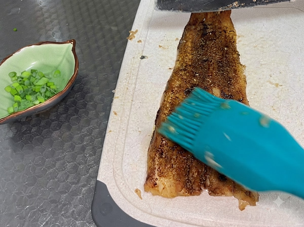 곱게 다진 쪽파가 담긴 초록색 작은 그릇 옆 흰색 도마 위에서 완전히 구워진 장어 필렛을 썰어내는 모습.