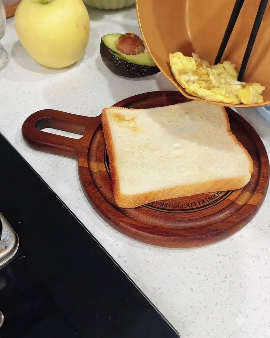 나무 도마 위 구운 하얀 식빵 조각 위에 구리빛 팬의 스크램블 에그를 젓가락으로 옮겨 담는 모습