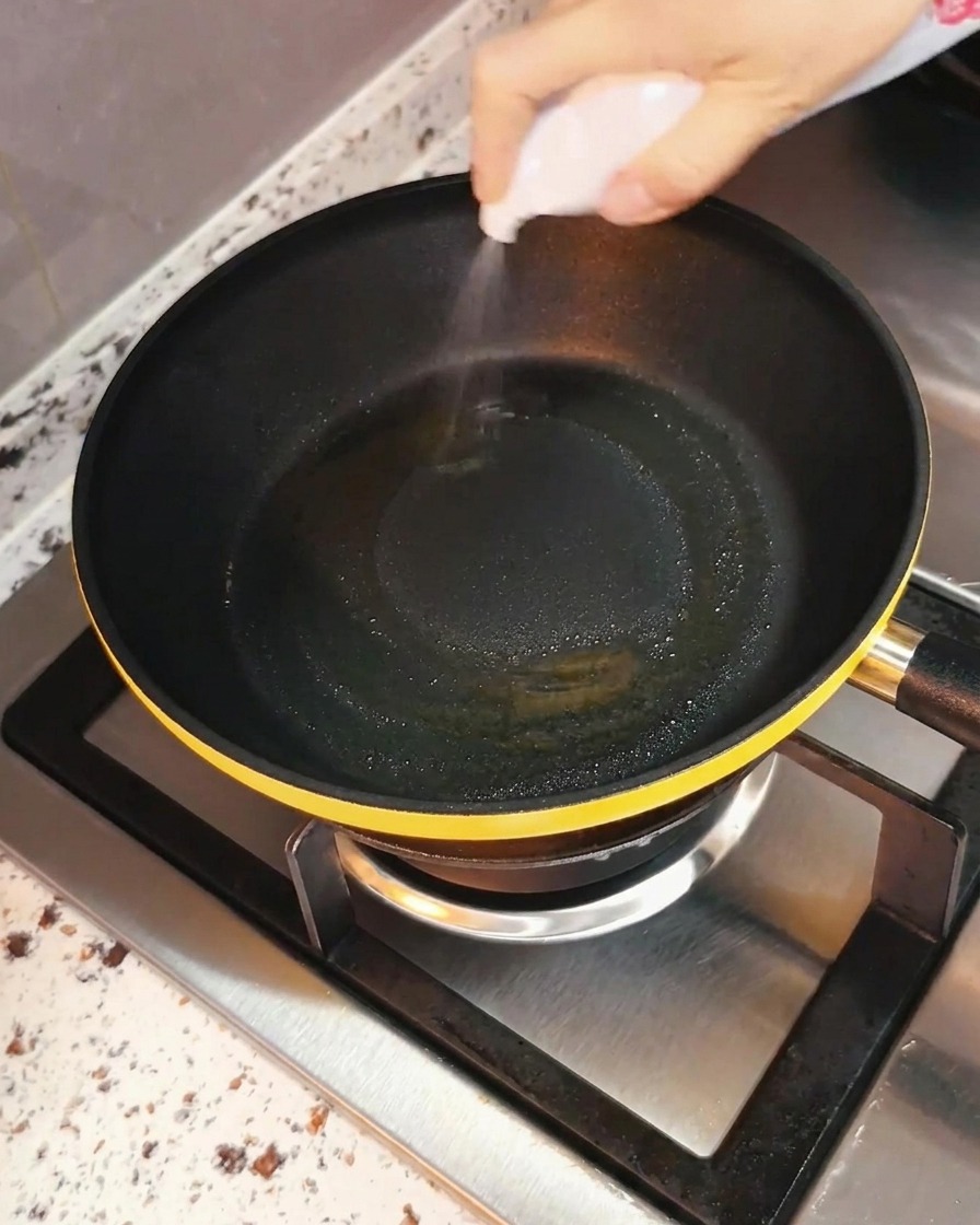 스토브 버너 위에 놓인 검은색 프라이팬에 손으로 식용유를 골고루 뿌리는 모습.