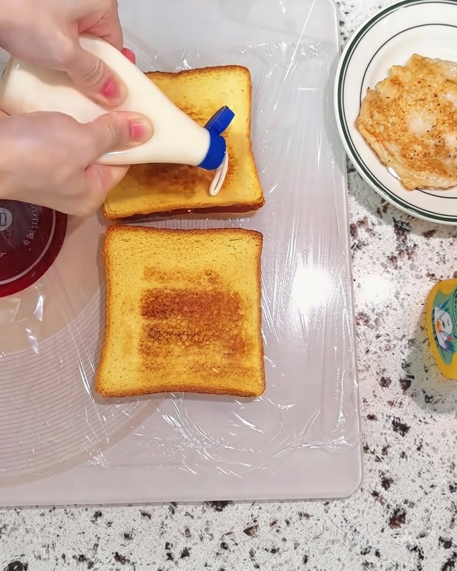 도마 위에 놓인 구운 빵 위에 소스 병에서 마요네즈를 짜는 모습.