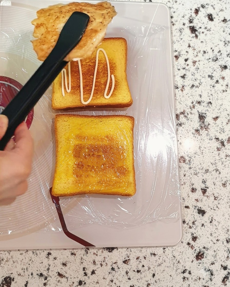 주방 집게로 조리된 닭가슴살을 소스가 발린 식빵 조각 위로 옮기는 모습.