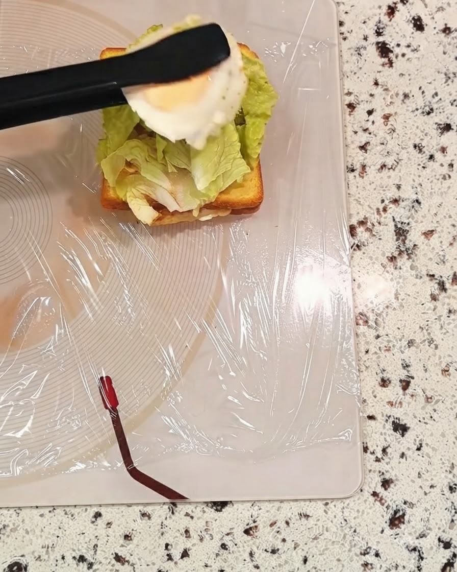 검은색 집게로 상추가 있는 샌드위치 층 위에 조리된 달걀을 집어 들고 있는 모습.