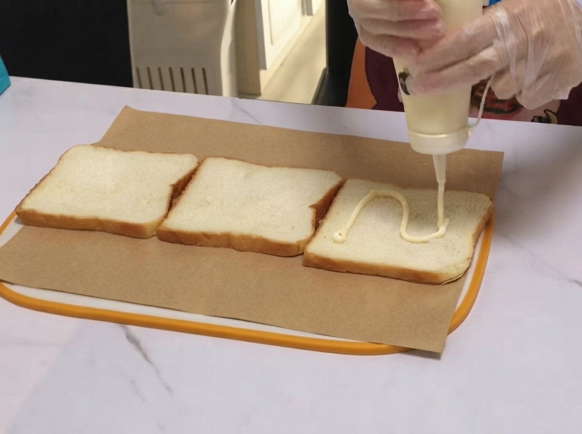 유산지 위에 놓인 식빵 조각에 화이트 소스가 물결 모양으로 짜여지는 모습.