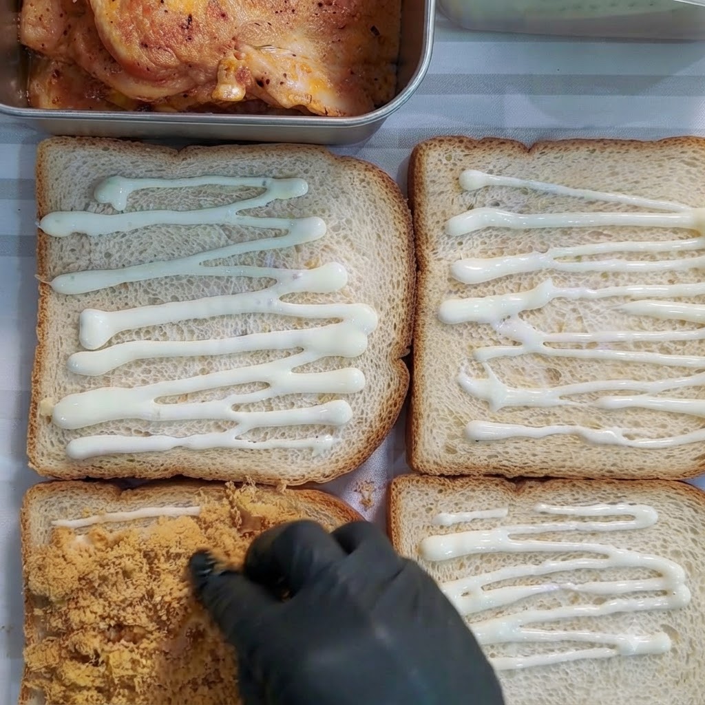 장갑을 낀 손이 드레싱이 발린 빵 조각 위에 황금빛 육송을 골고루 얹는 모습.