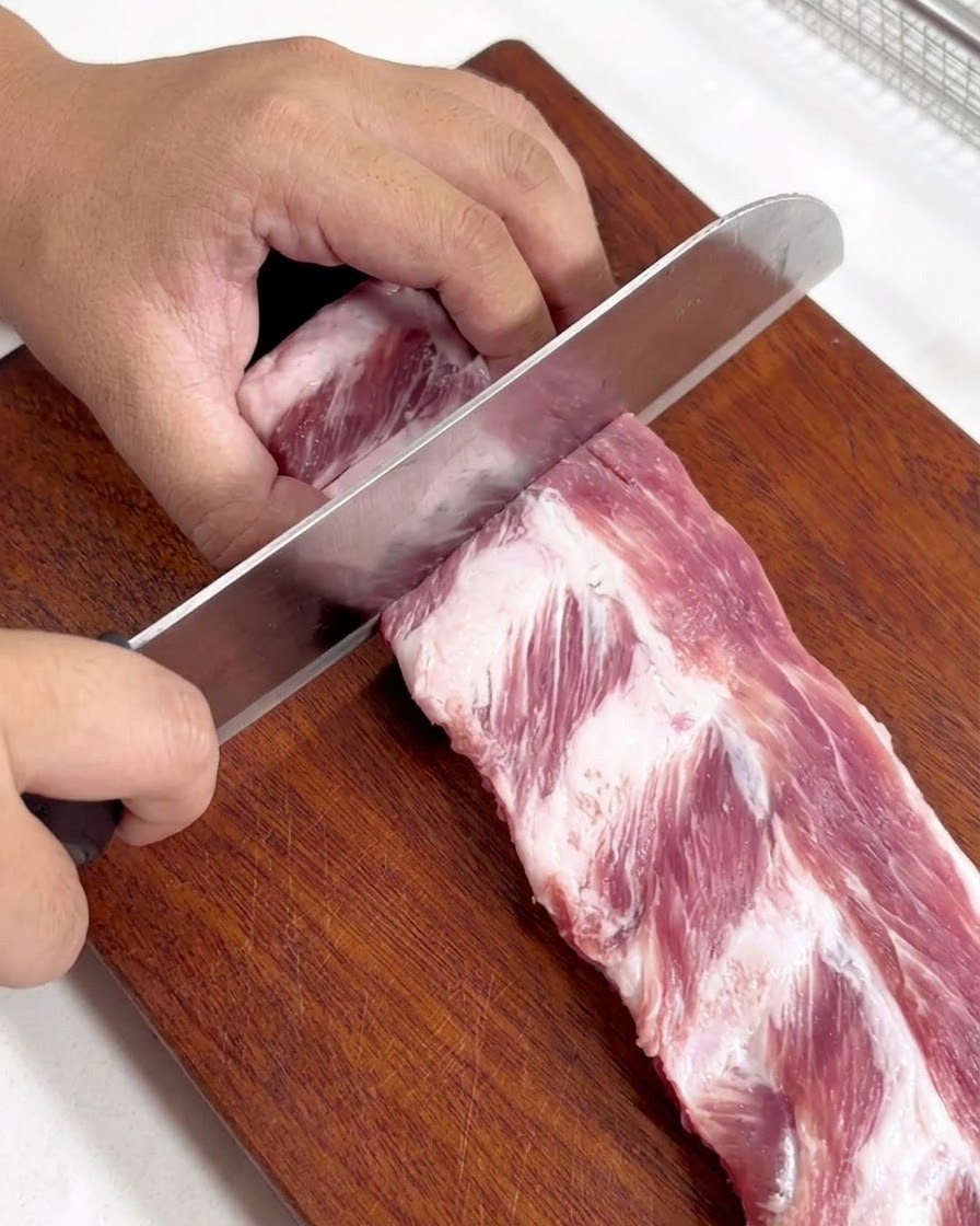 나무 도마 위에서 날카로운 칼로 생돼지갈비를 더 작은 한 입 크기로 자르는 모습.