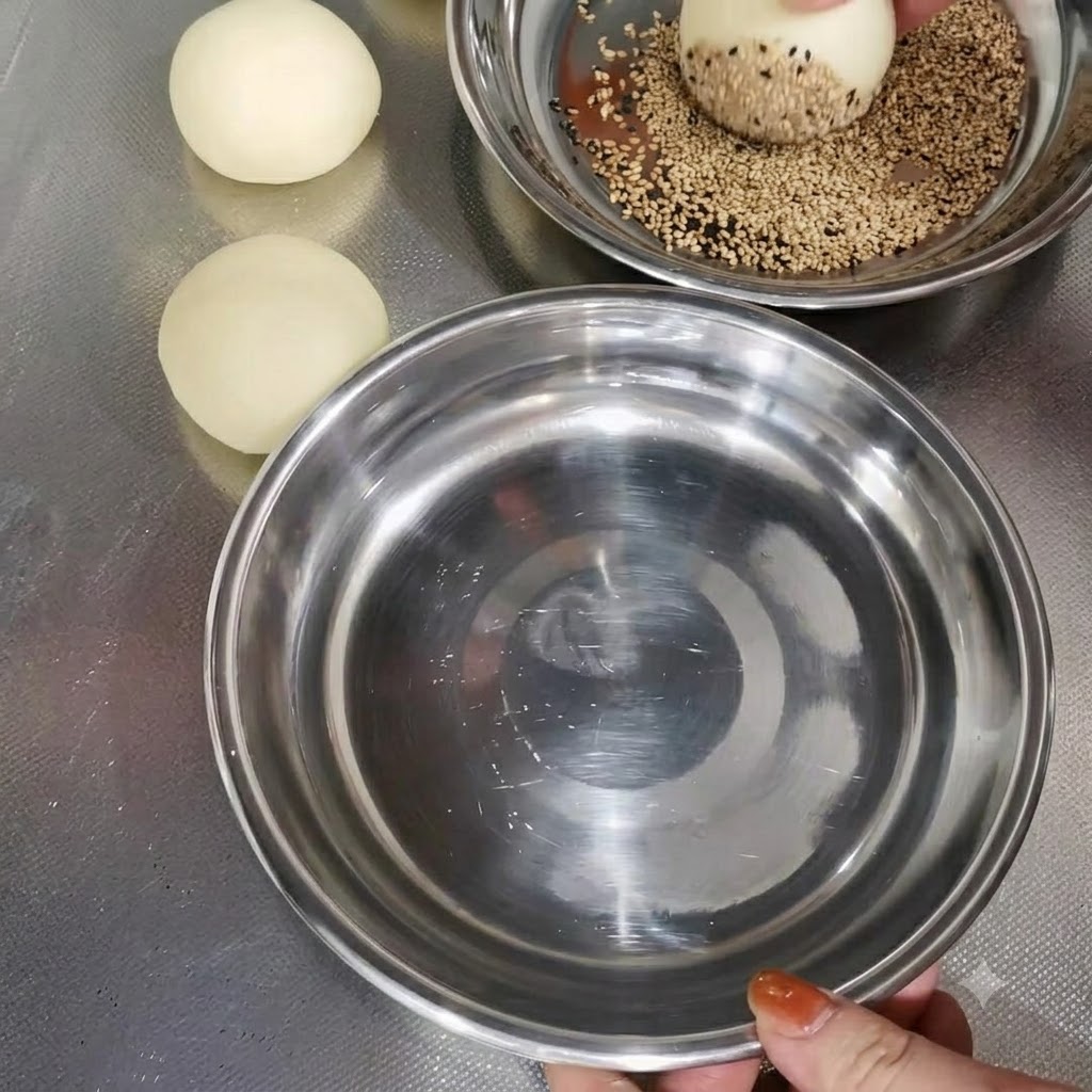 다른 반죽 덩어리들 옆 금속 그릇에서 참깨가 묻혀지고 있는 반죽.