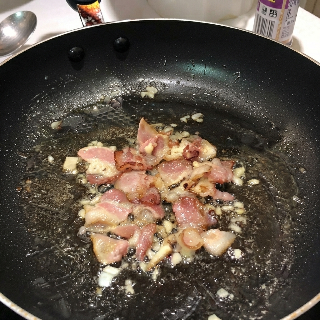 검은색 프라이팬 안에서 삶아진 펜네 파스타가 베이컨, 양파와 섞여 있는 모습.