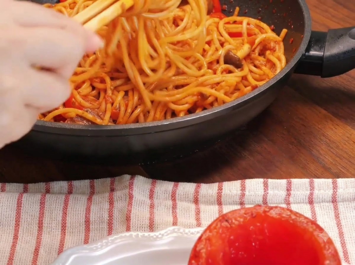 접시에 놓인 구운 토마토 그릇 옆에서 젓가락을 이용해 팬 안의 스파게티 면을 돌돌 말고 있는 클로즈업 샷.