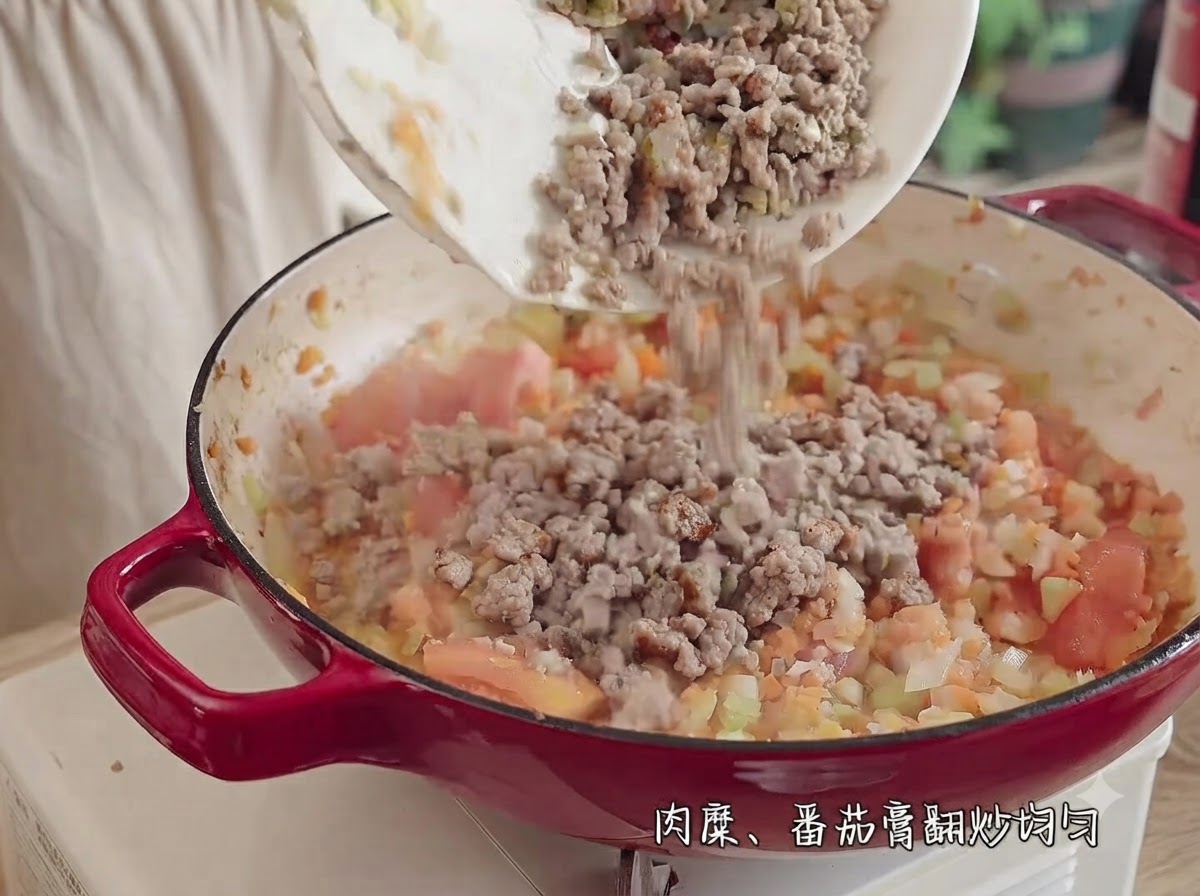 다진 토마토, 양파, 셀러리, 당근이 채워진 붉은 냄비에 하얀 접시의 간 고기를 붓는 모습.