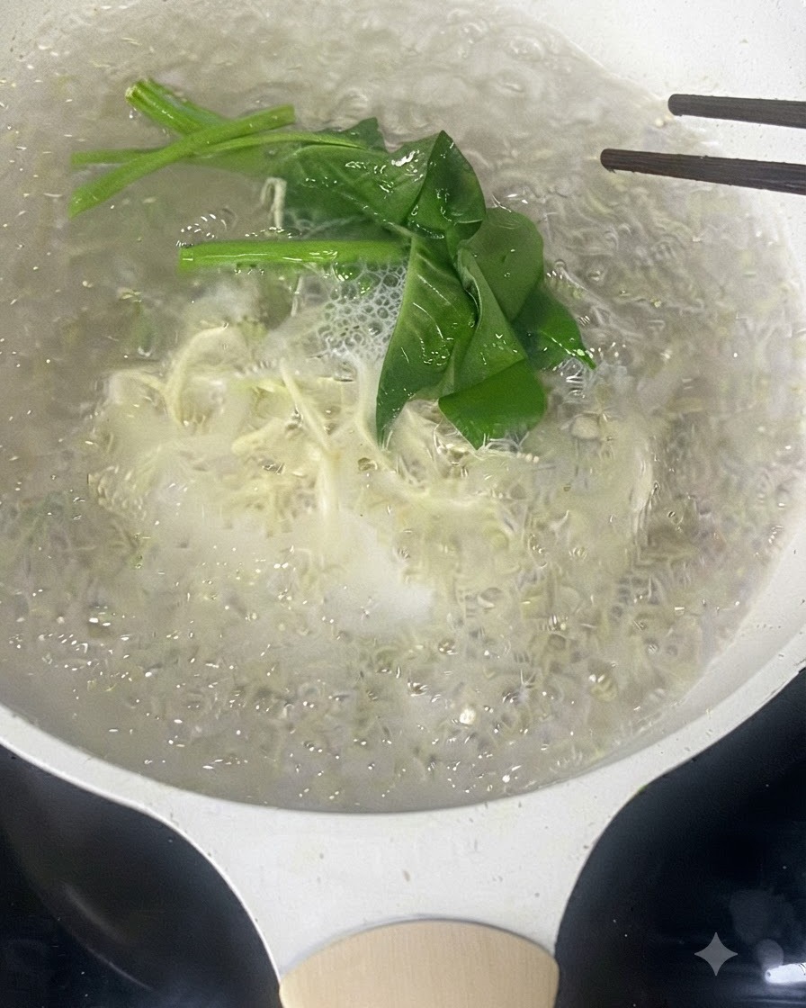 젓가락으로 끓는 물에 삶아지고 있는 가늘고 흰 국수와 초록색 잎채소를 젓는 모습.