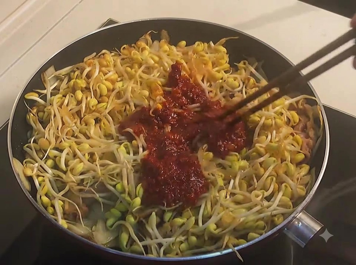 진한 붉은 양념장과 콩나물, 우삼겹을 젓가락으로 섞으며 볶아주는 모습.