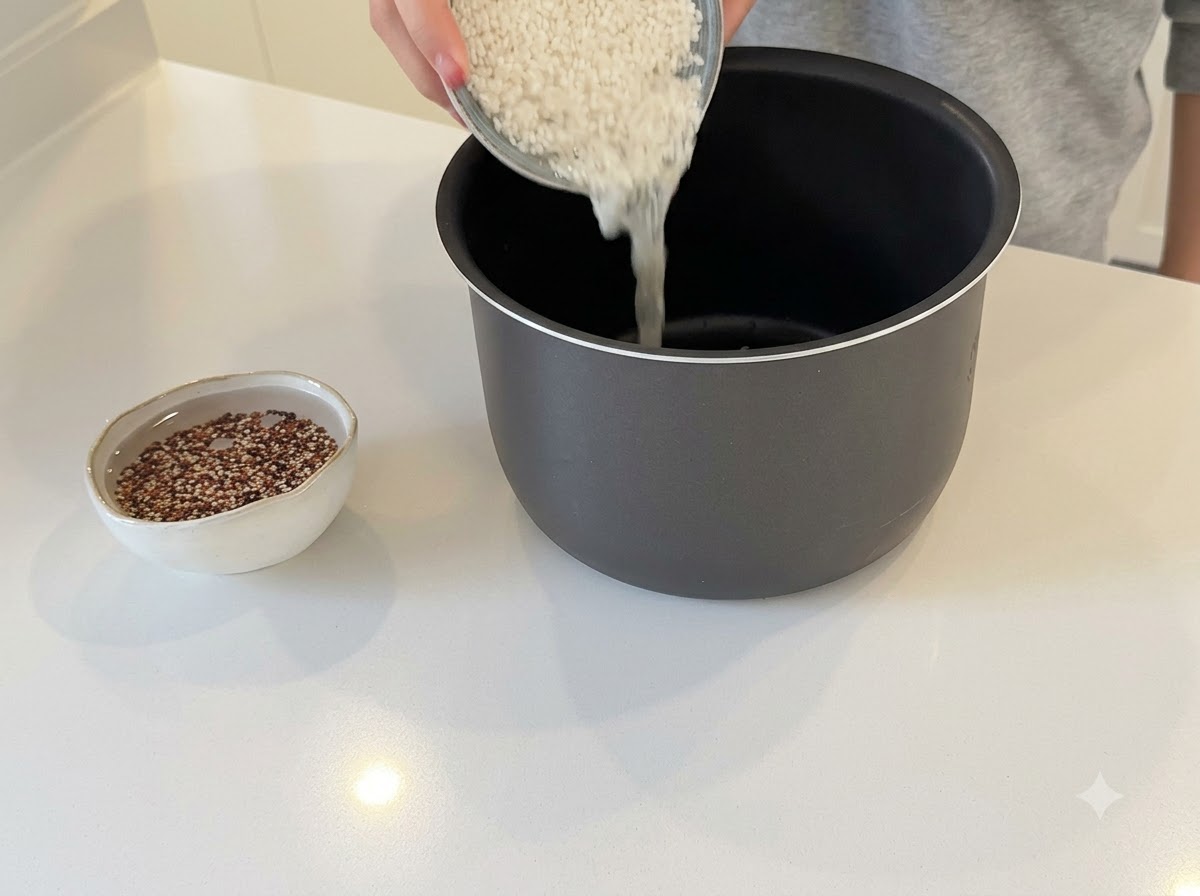 한 손으로 백미를 밥솥 내부에 붓고 있으며, 옆에는 퀴노아가 물에 불려지고 있는 작은 그릇이 놓여 있습니다.