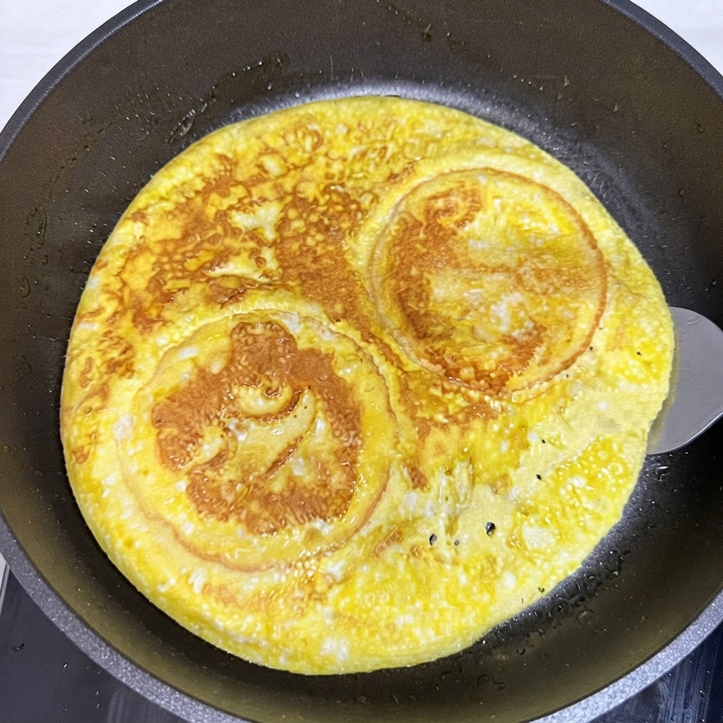 뒤집개로 프라이팬 안의 둥근 달걀 부침과 버거 빵 조합을 뒤집는 모습.