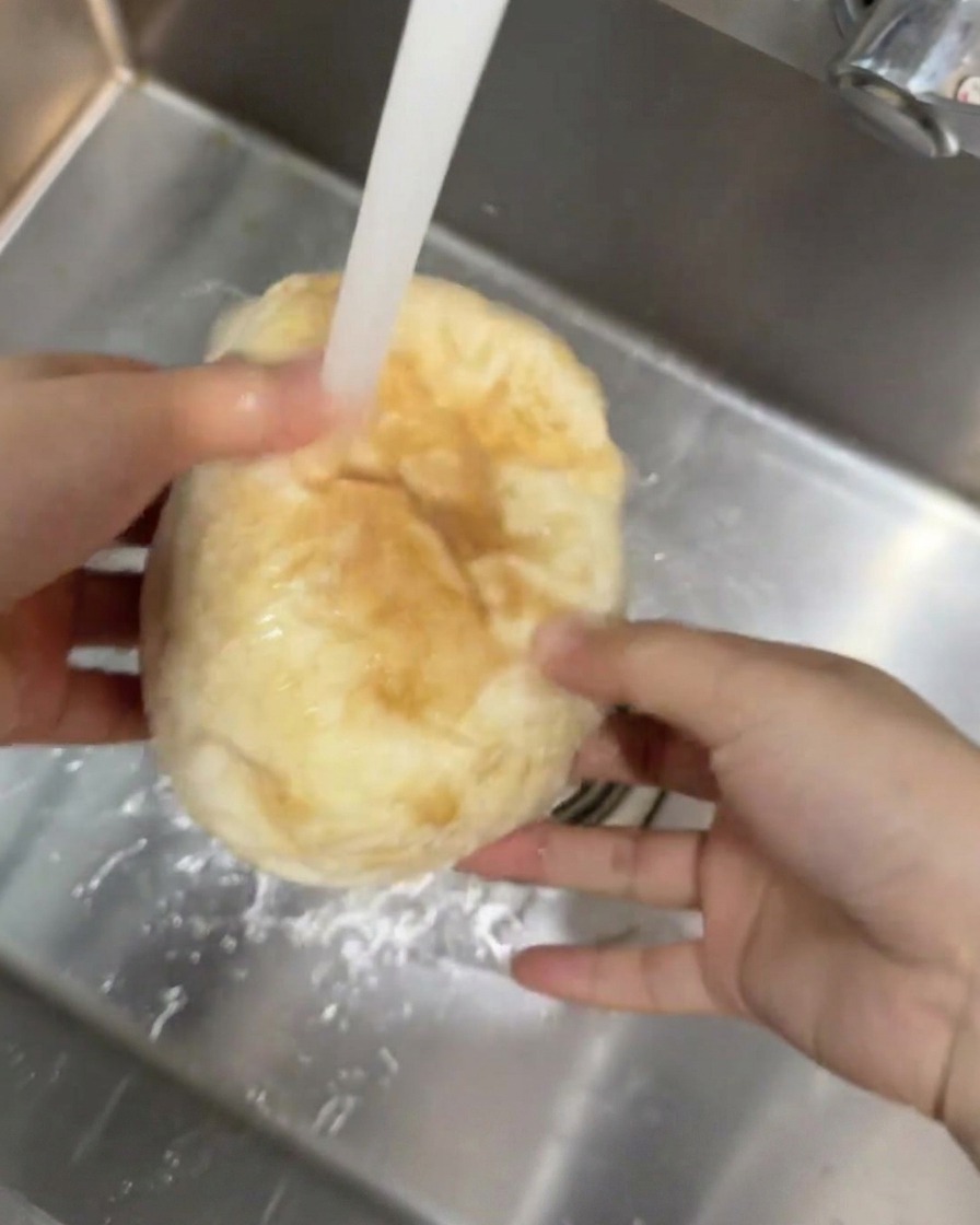 주방 싱크대 흐르는 수돗물 아래에서 둥근 치아바타 빵을 손으로 잡고 헹구는 모습.