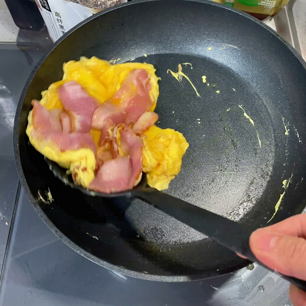 뒤집개로 익은 계란과 베이컨 층을 들어 올려 토스트한 잉글리시 머핀 절반 위에 올리는 모습.