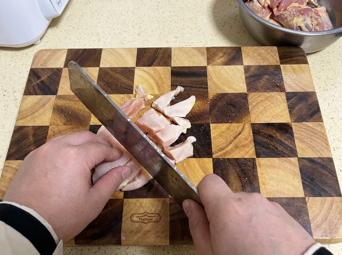 격자무늬 나무 도마 위에서 식도로 생닭 가슴살을 작게 썰고 있는 모습.