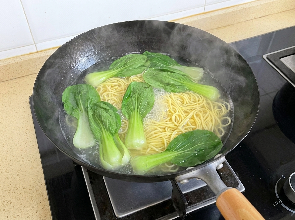 웍에서 끓고 있는 국수와 함께 신선한 청경채 잎이 끓는 물 위에 떠 있는 모습.