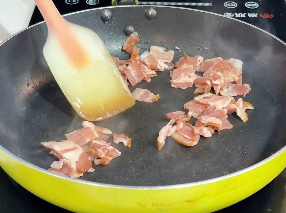팬 안에서 주걱으로 베이컨과 양파를 볶아내는 모습.