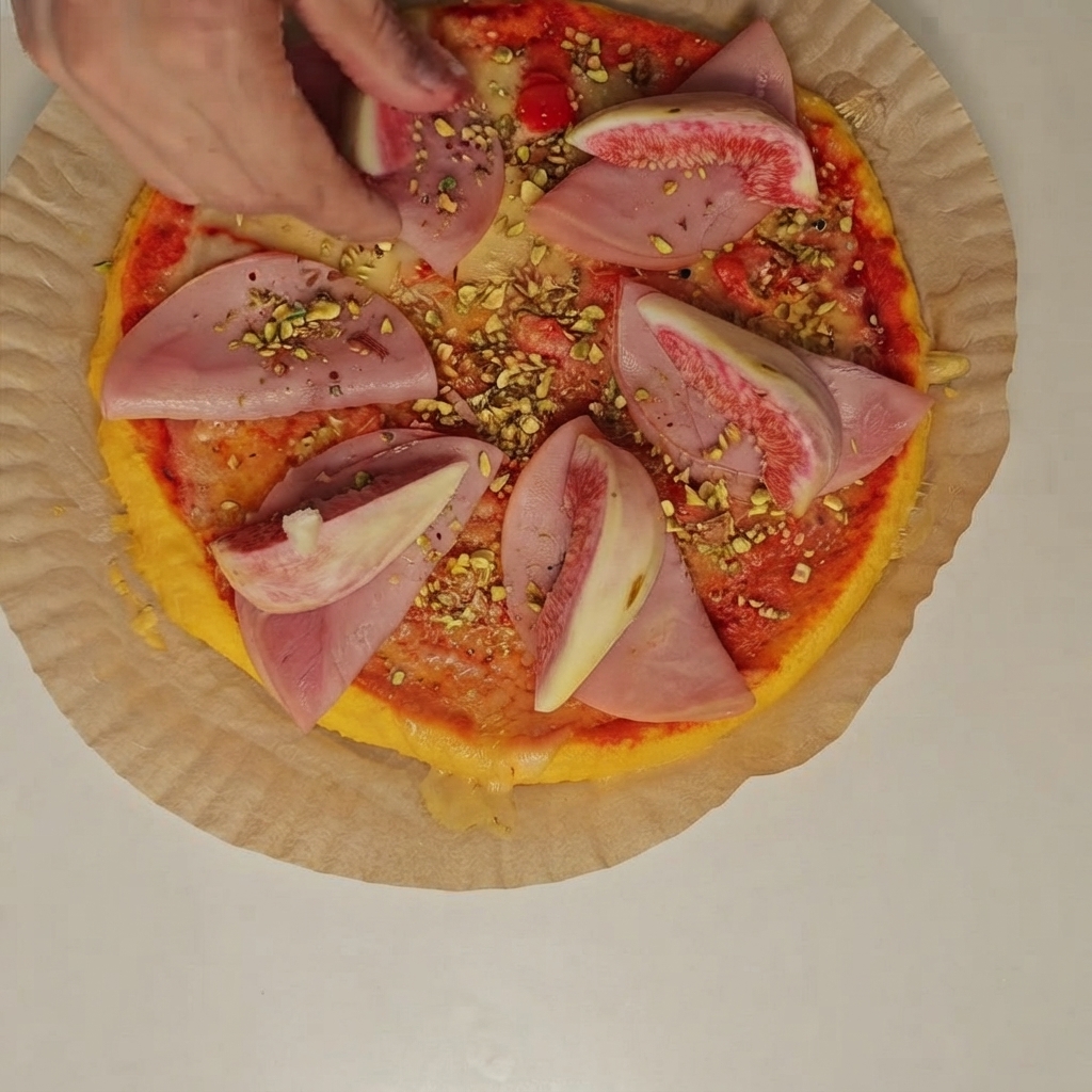 피자 위 햄 조각 위에 신선한 무화과 조각과 다진 피스타치오를 올리는 사람의 손.