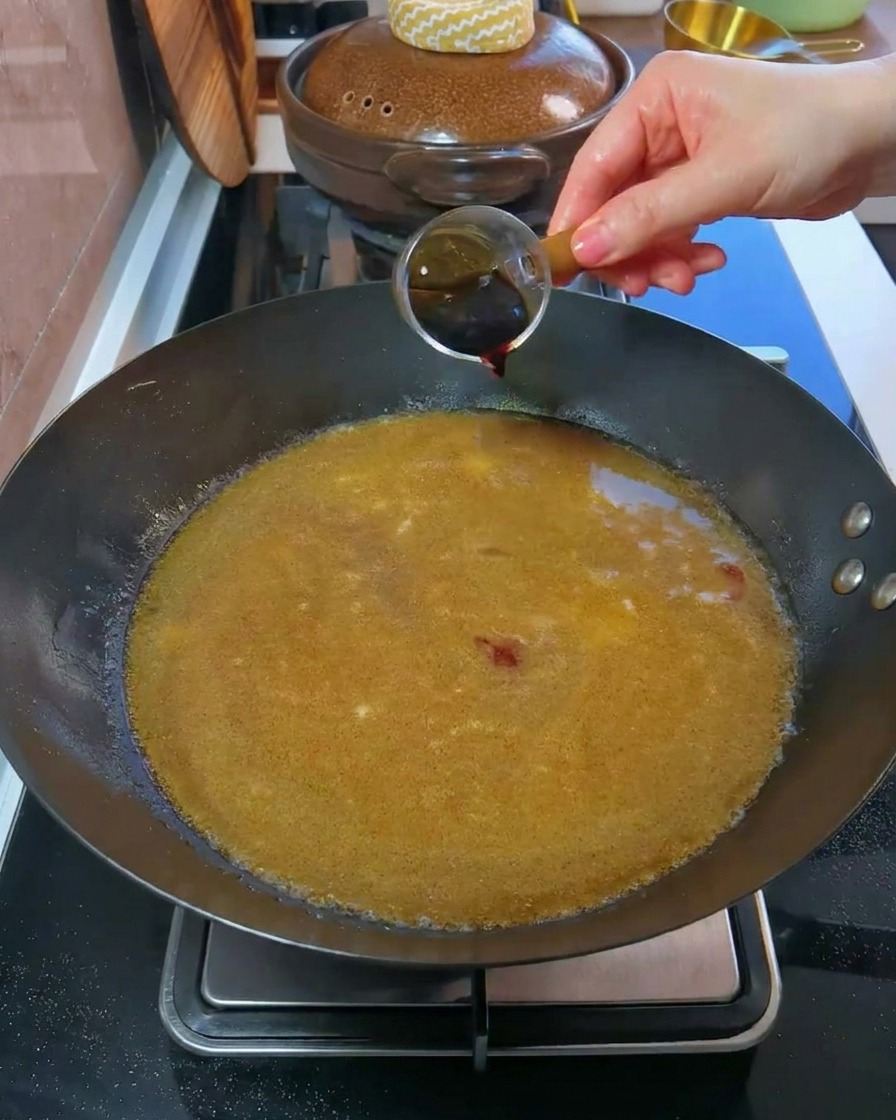 작은 계량컵에 담긴 진간장이 끓고 있는 육수가 가득 찬 웍으로 손을 통해 부어지는 모습.