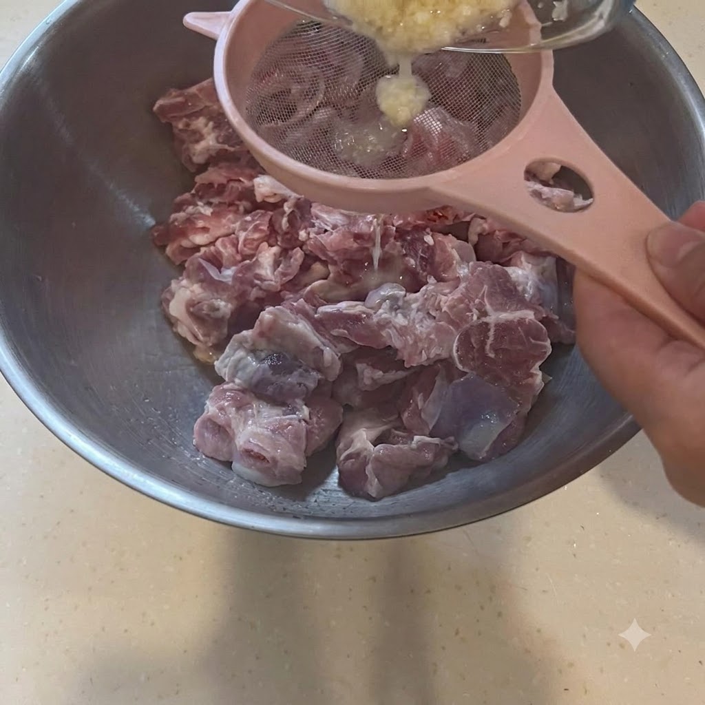 생돼지고기 조각이 담긴 큰 볼 위 분홍색 체에 마늘 페이스트를 거르는 모습.
