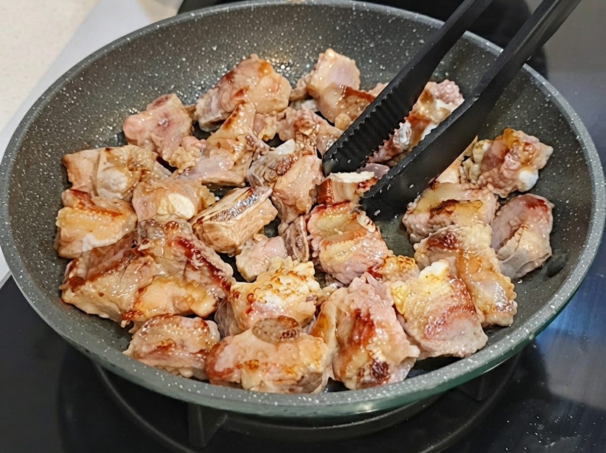 검은색 집게로 넌스틱 프라이팬 안의 돼지갈비를 뒤집는 모습.