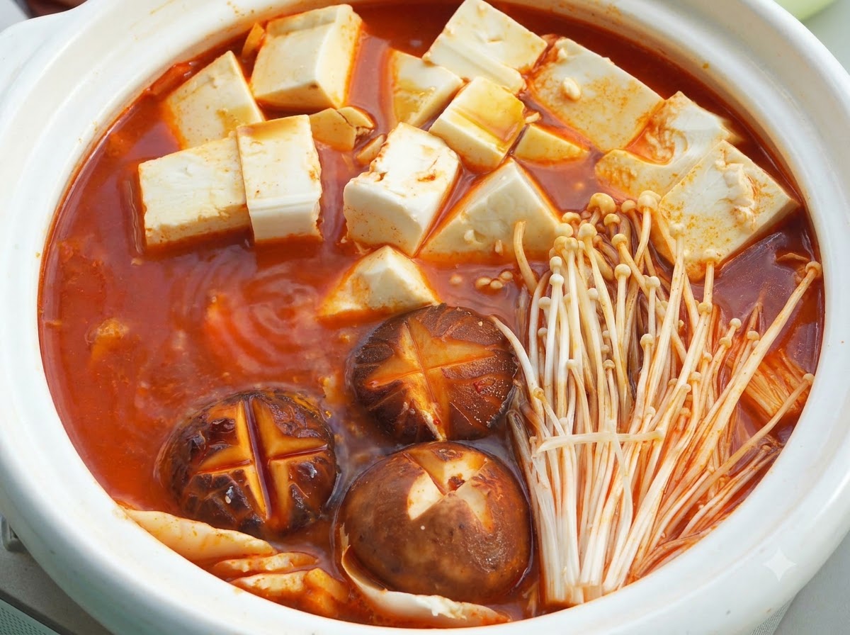 붉은 국물, 순두부, 십자 모양을 낸 표고버섯, 팽이버섯이 든 뚜껑 덮인 흰색 세라믹 냄비.
