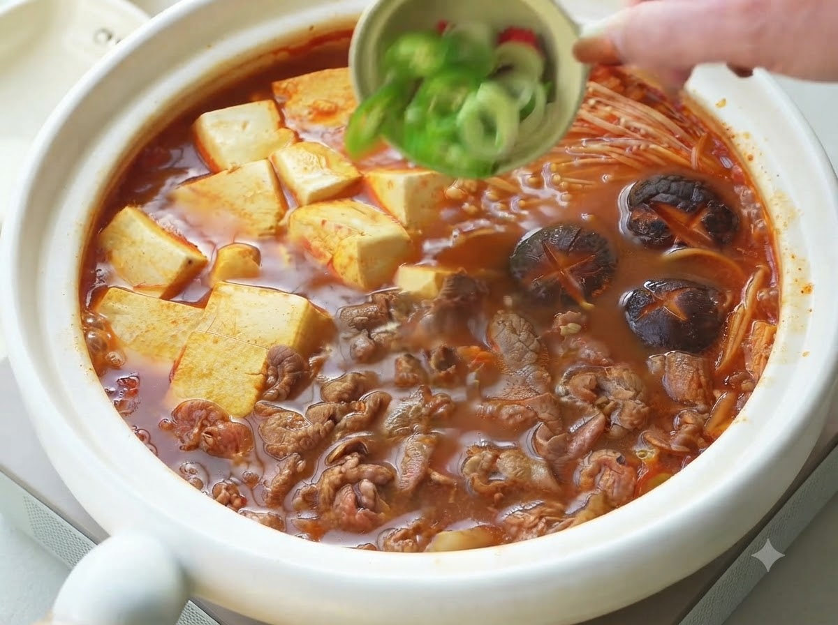 완성된 소고기 순두부 찌개 위에 작은 볼에 담긴 대파와 고추를 고명으로 뿌리는 모습.